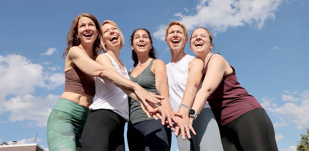 Fünf Frauen in Sportkleidung stehen draußen im Kreis, lachen und legen die Hände übereinander vor blauem Himmel.