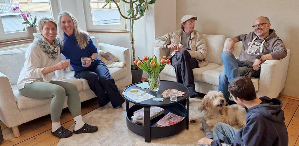 Heller, gemütlicher Yoga-Loungebereich im Yogaplus Studio Mainz: mehrere Erwachsene sitzen lachend auf Sofas, trinken Wasser und unterhalten sich; ein Junge spielt mit einem kleinen Wuschelhund am Boden.
