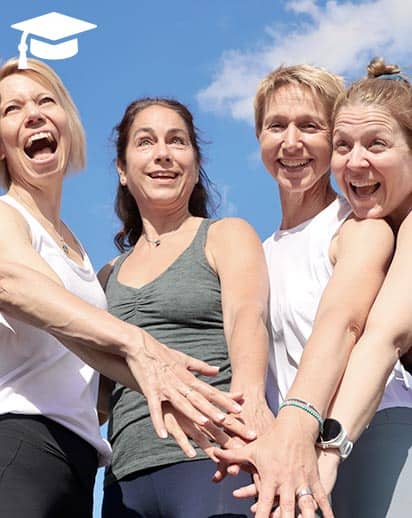 Fünf fröhliche Dozentinnen der Yogalehrerausbildung 300h bei Yogaplus stehen im Freien und legen motiviert die Hände übereinander vor blauem Himmel.