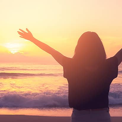 Frau am Strand mit ausgebreiteten Armen beim Sonnenaufgang, Meer und Wellen im Hintergrund, symbolisiert Freiheit und Achtsamkeit.