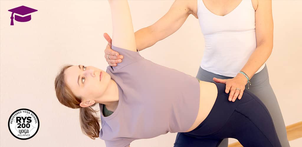 Eine Yogalehrerin korrigiert die Haltung einer Schülerin in einer seitlichen Yoga-Pose. Die Schülerin trägt ein lila Oberteil, die Lehrerin ein weißes. Oben links ist das lila Doktorhut-Symbol das Yogaplus Academy zu sehen, unten links das 'RYS 200'-Zertifizierungslogo der Yoga Alliance.