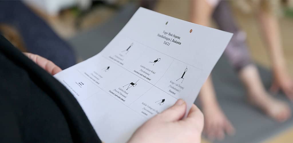 Seitlicher Blick über die Schulter einer Yogaschülerin auf ihren Anleitungsbogen mit schematischen Darstellungen von Asanas, während eine Schülerin im Hintergrund diese praktiziert