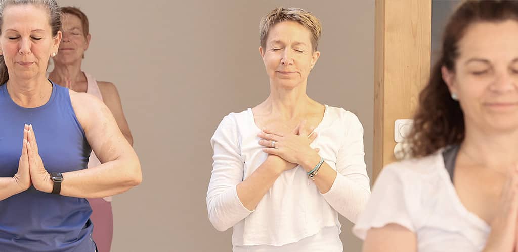 Yogalehrerin Susanne Weisheit steht während eines Kurses mit geschlossenen Augen und auf ihrer Brust zusammengelegten Händen zwischen ihren Yogaschülerinnen und meditiert mit ihnen zusammen