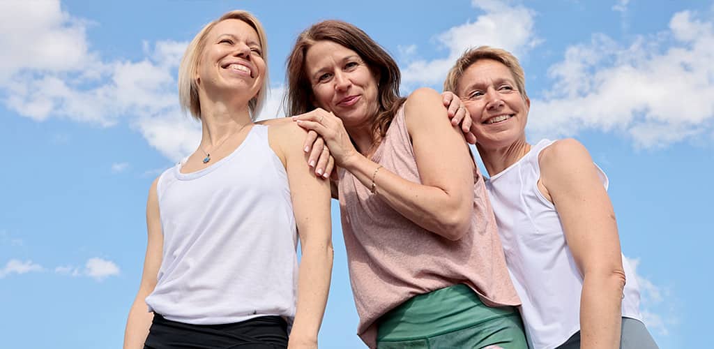 Drei Yogalehrerinnen Maja Sidebaeck, Sonja Schwarz-Niemeyer und Susanne Weisheit stehen in sportlichen schulterfreien Yogaoutfits vor blauen Sommerhimmel eng zusammen und lachen