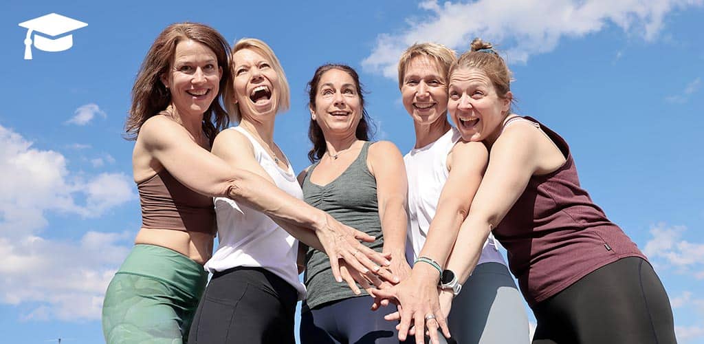Fünf fröhliche Dozentinnen der Yogalehrerausbildung 300h bei Yogaplus stehen im Freien und legen motiviert die Hände übereinander vor blauem Himmel.