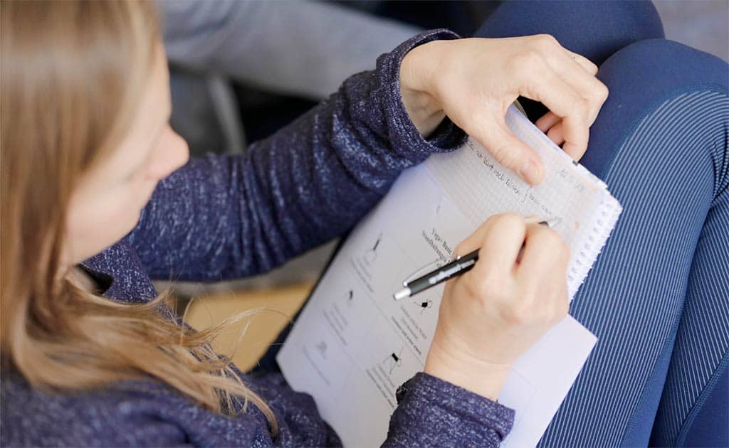 Teilnehmerin einer Yogaweiterbildung schreibt Notizen in ein Heft, auf dem ein Übungsblatt mit Yoga-Illustrationen liegt.