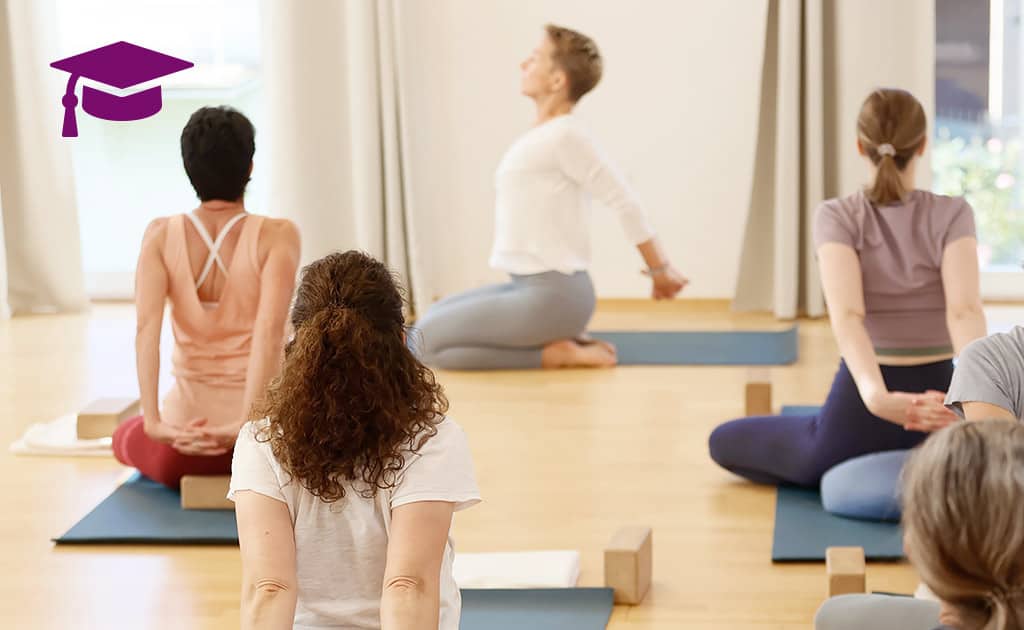 Yogagruppe sitzt auf Matten im Schneidersitz und verschränkt hinter dem Rücken die Arme, während Yogalehrerin Susanne Weisheit die Dehnung anleitet.