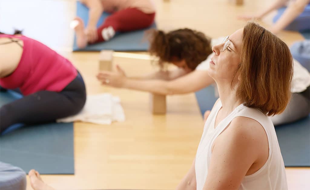 Gruppe von Menschen auf Yogamatten, eine Frau im Vordergrund sitzt mit geschlossenen Augen aufrecht, während andere sich nach vorne beugen.