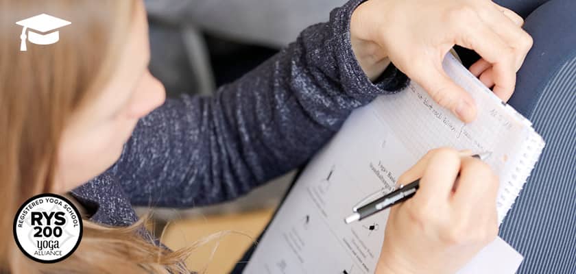 Teilnehmerin einer Yogaweiterbildung schreibt Notizen in ein Heft, auf dem ein Übungsblatt mit Yoga-Illustrationen liegt.