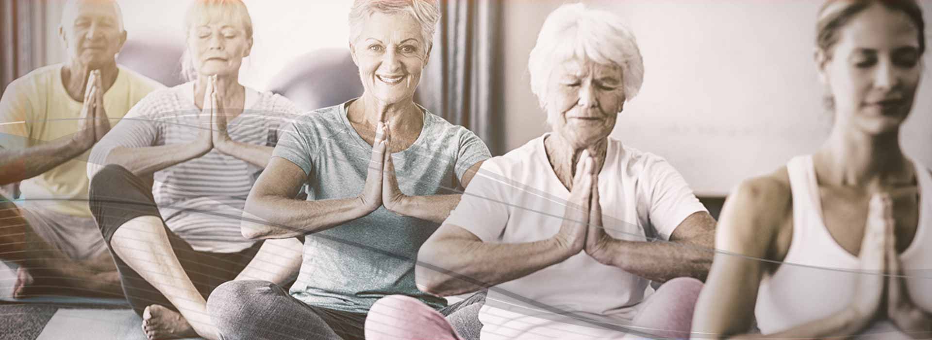 Eine Gruppe weißhaariger Senioren sitzen lächelnd zusammen mit ihrer jüngeren Yogalehrerin im Schneidersitz und vor der Brust gefalteten Händen auf ihren Yogamatten und meditieren