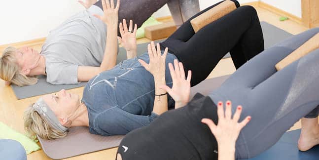 Drei Yogaschülerinnen üben auf ihren Yogamatten die Schulterbrücke und sichern ihre Haltung durch zwischen den Knien gepressten Yogablöcken