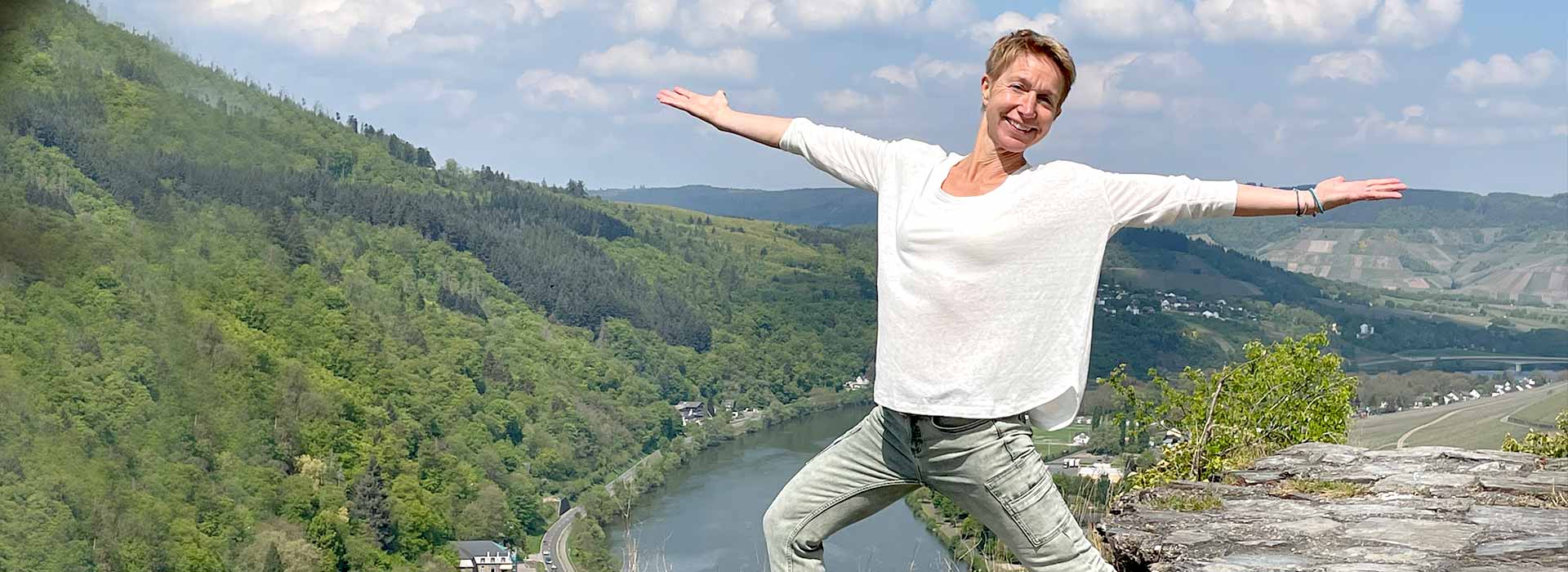 Yogalehrerin Susanne Weisheit steht in blauer Jeans und hellem Langarmshirt mit weit ausgereiteten Armen auf einem Mauerrest der Burgruine Grevenstein über Traben-Trabach mit Blick ins Moseltal und lacht in die Kamera