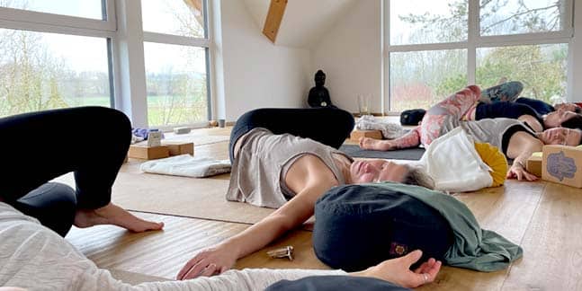Die Teilnehmer des Yogaplus-Mini-Retreats im hellen Yogaraum von Jordan's Untermühle in seitlicher Drehung und mit ausgestreckten Armen auf dem Rücken und entspannen mit geschlossenen Augen auf ihren Yogamatten.