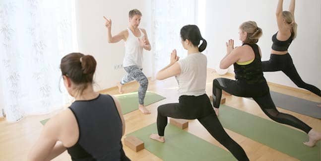 Yogalehrerin Susanne Weisheit steht in ihrem Studioraum vor einer Gruppe von vier Yogaschülerinnen und übt zusammen mit Innehalten die Asana 'tiefer Ausfallschritt'