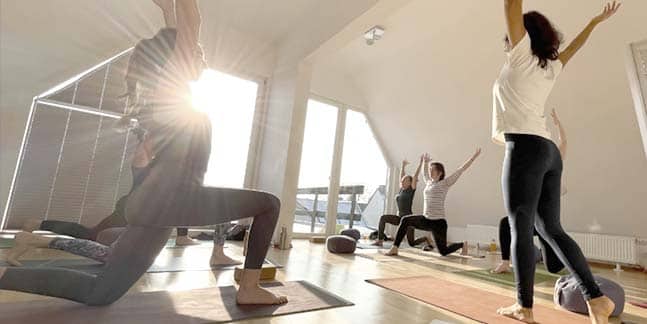 Eine Gruppe von Absolventen der Yogaplus Yogalehrerinnenausbildung 2022 praktiziert im hellen sonnendurchfluteten Dachraum vor der verglasten Stirnseite auf ihren Yogamatten die Asana 'Krieger 1'