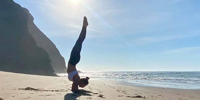 Yogalehrerin Claudia Fleiner praktiziert die Asana 'Pincha-Mayurasana' im Gegenlicht der Sonne am Sandstrand bei wolkenlosem blauem Himmel und Blick auf das ruhige Meer