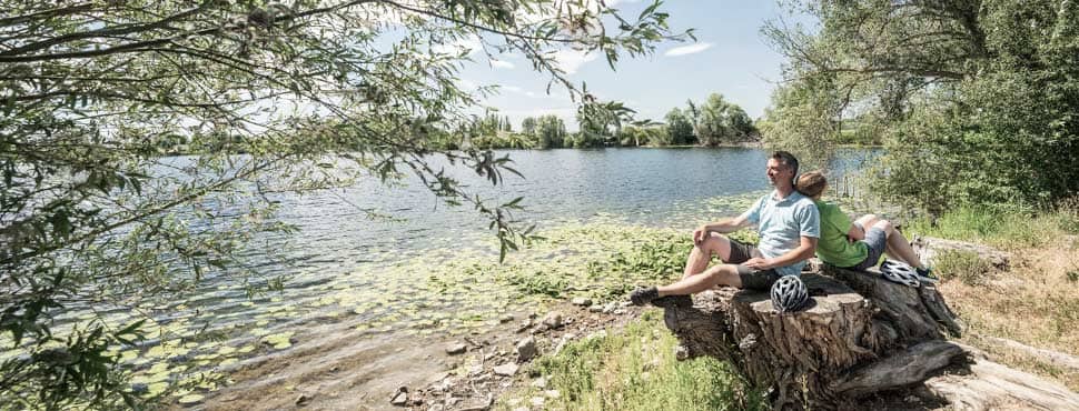 Junges Paar in kurzen Hosen und Wanderoutfit sitzt Rücken an Rücken bei sonnig-blauem Himmel entspannt auf einem großen Baumstumpf in einer Auenlandschaft am Rheinufer und macht Pause