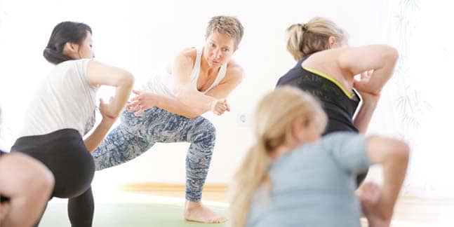 Yogalehrerin Susanne Weisheit gibt in einer hellblau-gemusterten Yogaleggings mit schulterlosem weißen Top in tiefem Ausfallschritt auf einer grünen Yogamatte einer Gruppe von Yogaschülern Anweisungen, während sie die Asana 'Tadasana' üben