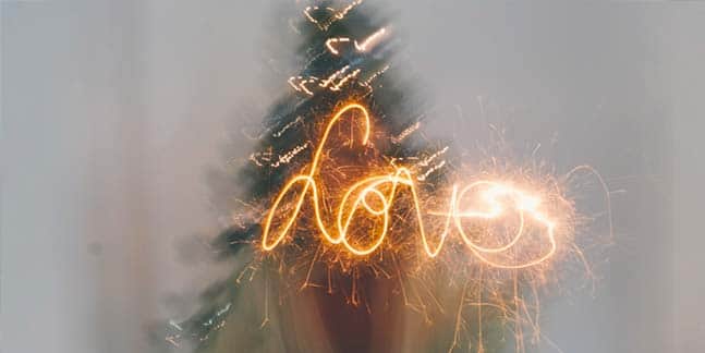 Leuchtspur einer Wunderkerze beschreibt den Schriftzug LOVE vor unscharf abgebildeten dekorierten Weihnachtsbaum