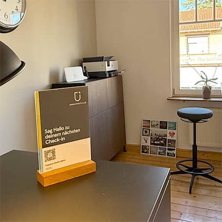 Der helle Empfangsbereich vom Yogaplus Studio Mainz mit Stehtisch, Hockern, Wandlampe, Infomaterial und großem Fenster; moderne, einladende Studioatmosphäre zum Wohlfühlen.