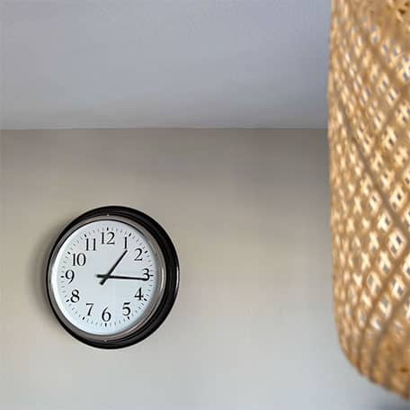 Detail Empfangsbereich vom Yogaplus Studio Mainz: runde Wanduhr an hellgrauer Wand, im Vordergrund eine große geflochtene Hängelampe.