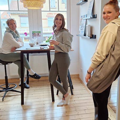 Drei Frauen im Empfangsbereich vom Yogaplus Studio Mainz: zwei sitzen am hohen Fenster­tisch mit Getränken, eine steht mit Yogamatte über der Schulter; helle Holzboden, Regale und geflochtene Lampe.