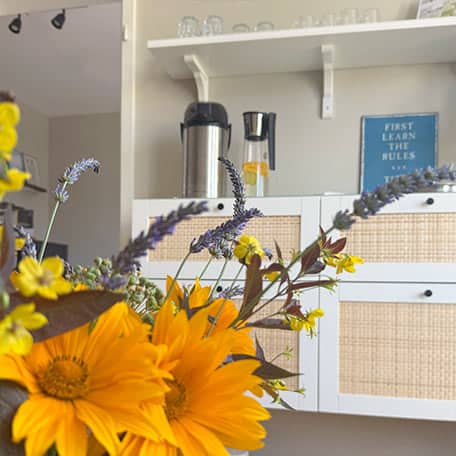 Nahaufnahme eines bunten Blumenstraußes mit gelben Sonnenblumen und Lavendel; im unscharfen Hintergrund eine helle Studioküche mit Thermoskannen und Schild „First learn the rules“.