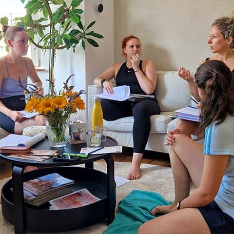 Gruppe von fünf Frauen in der hellen Studio-Lounge des Yogaplus Studio Mainz diskutiert sitzend auf Sofa und Boden; Notizblöcke und Getränke auf dem runden Couchtisch mit Blumen, große Fenster und Zimmerpflanze im Hintergrund.