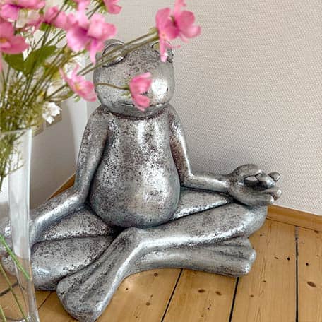 Silberne Frosch-Skulptur in Meditationspose neben rosa Blumen in Glasvasen auf Holzboden, vor heller Wand im Treppenhaus vom Yogaplus Studio Mainz