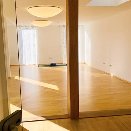 Der große heller Yogaraum im Yogaplus Studio Mainz mit Holzfußboden und runden Deckenleuchten; durch Glastür sichtbar, mittig eine Matte mit Bolster vor bodentiefen Fenstern.