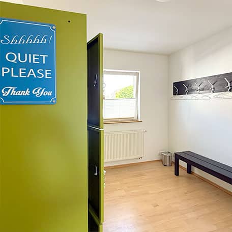 Heller Umkleideraum im Yogaplus Studio Mainz mit grünem Spind und blauem „Quiet Please“-Schild, langer Bank, Kleiderhakenleiste und Fenster.