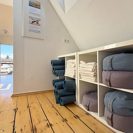 Offenes Yoga-Studiobereich mit Holzfußboden; Regal mit aufgerollten Yogamatten, weißen Handtüchern, Meditationskissen und Korkblöcken; im Hintergrund helle Fenster zur Dachterrasse.