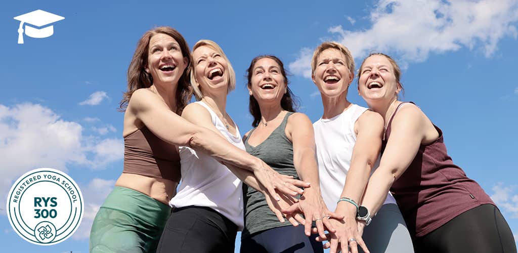 Fünf fröhliche Dozentinnen der Yogalehrerausbildung 300h bei Yogaplus stehen im Freien und legen motiviert die Hände übereinander vor blauem Himmel.