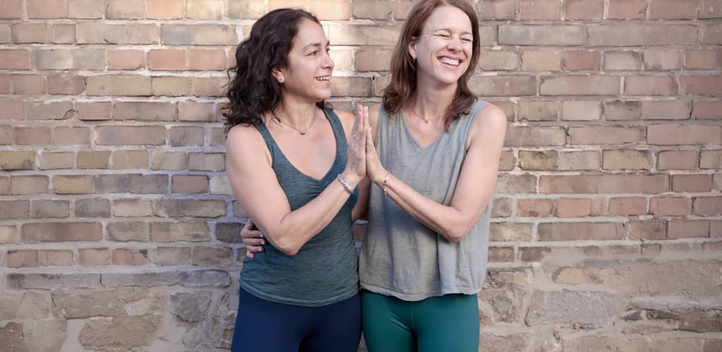 Zwei Yogalehrerinnen in Sportkleidung stehen lachend vor einer Backsteinmauer und geben sich freundschaftlich ein High-Five.