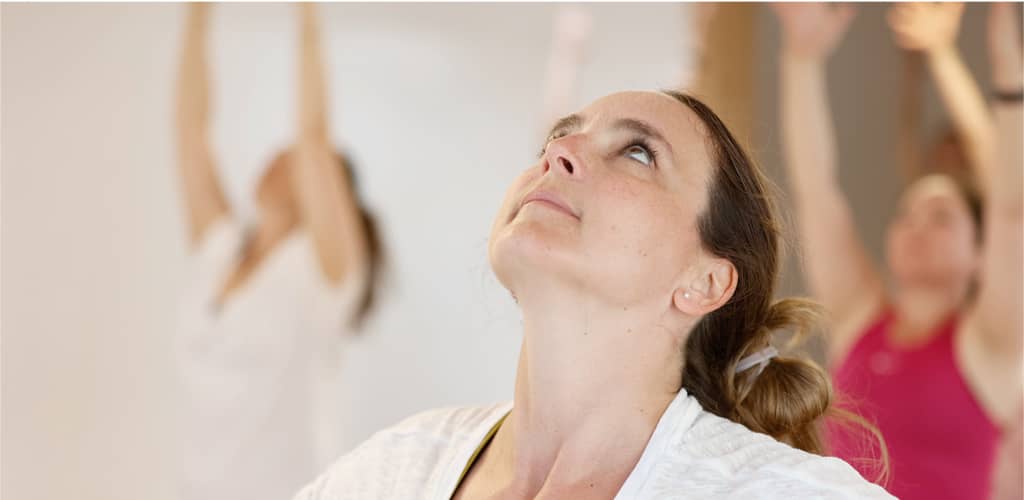 Frau im Yoga-Kurs blickt nach oben, während im Hintergrund weitere Teilnehmende die Arme heben.