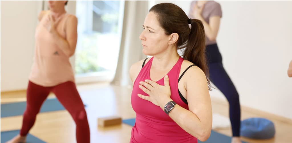 Frau in pinkem Sporttop steht mit Hand auf dem Herzen im Yoga-Kurs, im Hintergrund weitere Teilnehmende in Bewegung.