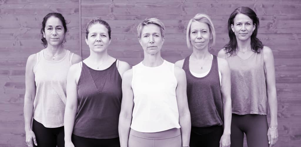 Fünf Yogalehrerinnen der Yogaplus Academy stehen in Sportkleidung vor einer Holzwand und blicken ruhig in die Kamera.