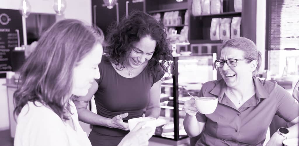 Drei Frauen sitzen lachend in einem Café zusammen und trinken aus Tassen; die Szene ist violett getönt.