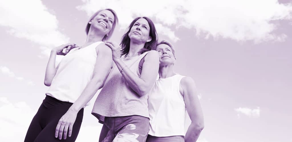 Drei Frauen in Sportkleidung stehen eng zusammen und blicken lächelnd in den Himmel; die Aufnahme ist violett getönt.