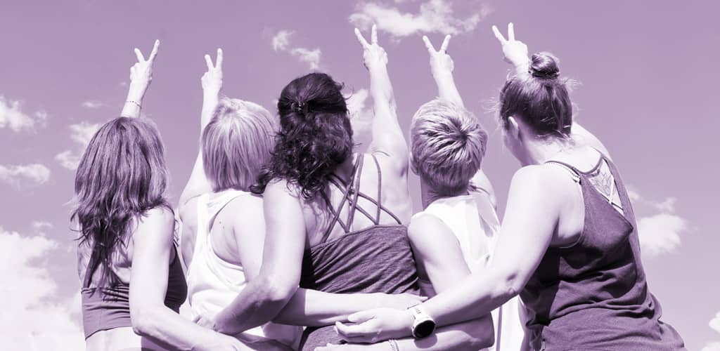 Fünf Frauen stehen mit den Armen umeinander und zeigen Friedenszeichen in den Himmel; die Aufnahme ist violett getönt.