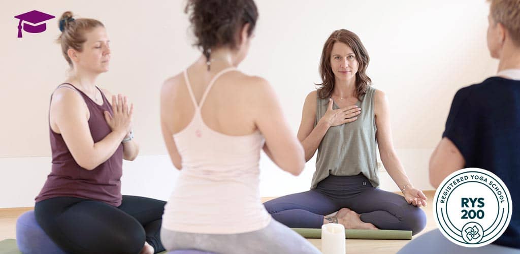 Yoga-Dozentin Sonja Schwarz-Niemeyer sitzt lächelnd im Kreis, Hand auf dem Herzen, im achtsamen Austausch mit Kursteilnehmerinnen. RYS-200-Zertifizierungssiegel eingeblendet.