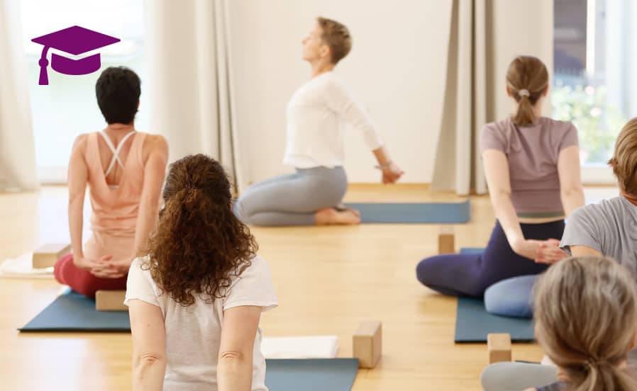 Yogagruppe sitzt auf Matten im Schneidersitz und verschränkt hinter dem Rücken die Arme, während Yogalehrerin Susanne Weisheit die Dehnung anleitet.