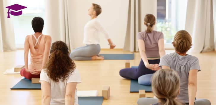 Yogagruppe sitzt auf Matten im Schneidersitz und verschränkt hinter dem Rücken die Arme, während Yogalehrerin Susanne Weisheit die Dehnung anleitet.