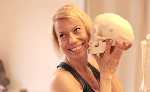 Lächelnde Yogalehrerin Maja Sidebäck hält ein anatomisches Schädelmodell an ihre Wange, helle Innenraumszene mit Lern- oder Therapiebezug.