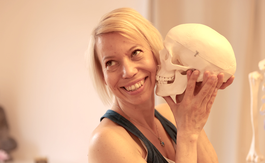 Lächelnde Yogalehrerin Maja Sidebäck hält ein anatomisches Schädelmodell an ihre Wange, helle Innenraumszene mit Lern- oder Therapiebezug.