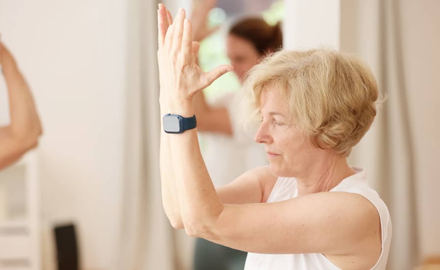 Eine ältere Frau mit blondem Haar und weißem Top praktiziert Yoga in einem hellen Raum. Sie konzentriert sich auf eine Übung mit erhobenen Armen, während andere Teilnehmer im Hintergrund zu sehen sind.
