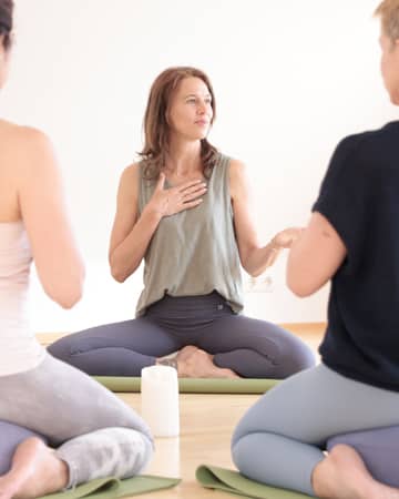 Kleine Yoga-Gruppe sitzt im Kreis; eine Frau spricht mit Hand auf dem Herzen, die anderen hören achtsam zu.