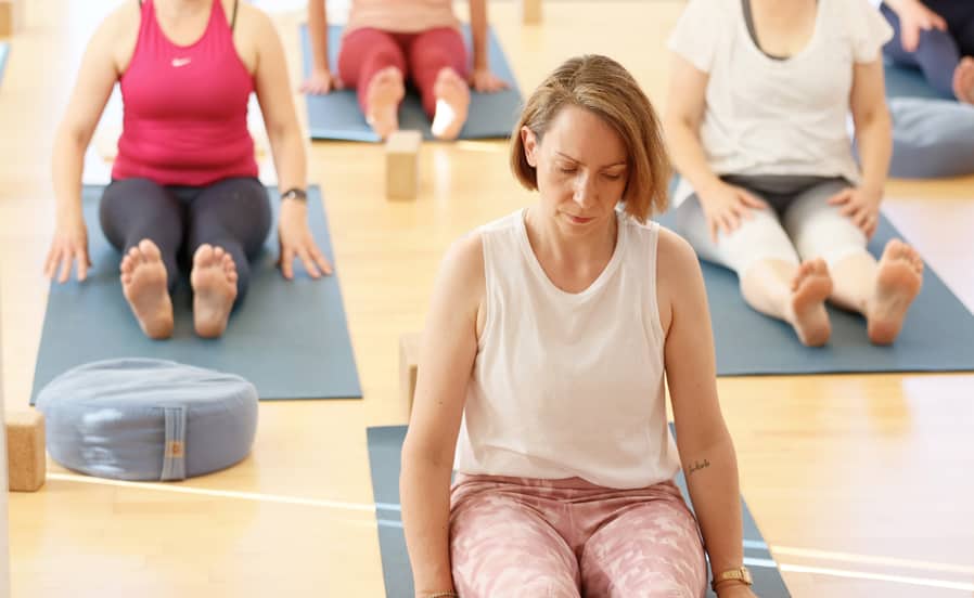 Eine Gruppe von Absolventen der Yogalehrer:innenausbildung bei Yogaplus sitzt auf Yogamatten in einer Meditationshaltung. Die Teilnehmer wirken konzentriert und entspannt, und Yoga-Props wie Blöcke und Kissen sind im Raum verteilt.