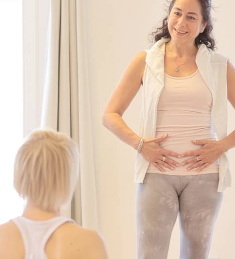 Yogalehrerin Nathalie Butscher steht erklärend mit einer auf ihrem Bauch platzierten Hand in sandfarbenem Oberteil und grauer Sportleggings während einer Seminarstunde vor ihren auf dem Boden sitzenden Teilnehmerinnen