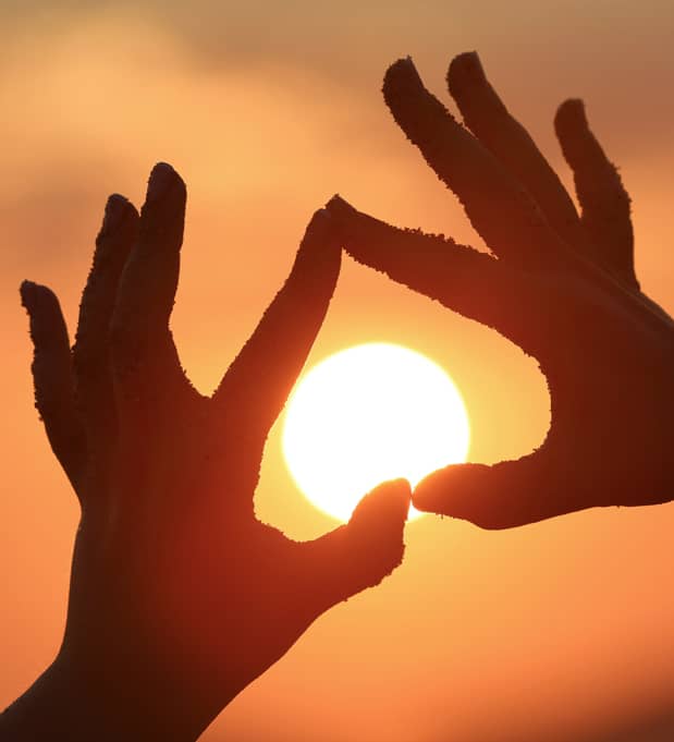 Silhouetten von Händen formen ein Herz vor der tief stehenden Sonne am Abendhimmel.